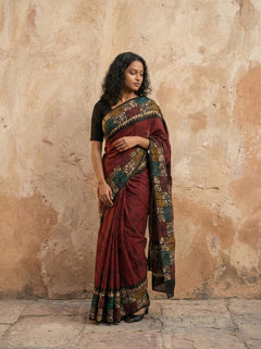 Woman wearing a traditional maroon saree with a patterned border against a textured wall.
