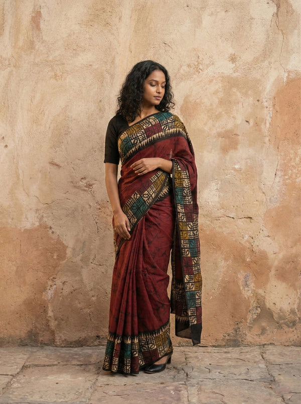 Woman wearing a traditional maroon saree with a patterned border against a textured wall.