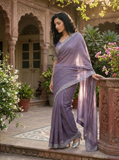 Woman in a lavender saree standing in an outdoor setting with architectural elements and plants.