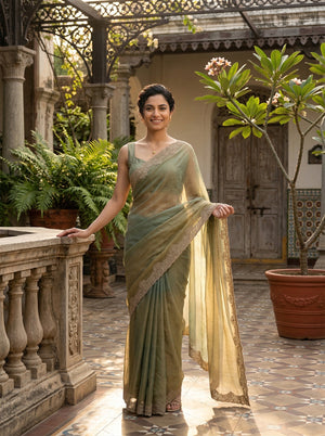Woman in a green saree standing in an outdoor setting with plants and architectural elements.