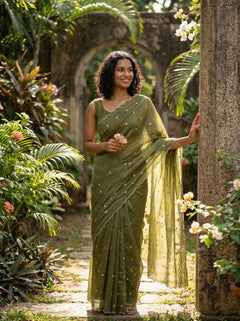 Woman in a green saree standing in a garden with plants and flowers around her.