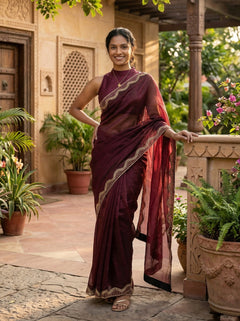 Woman in a maroon saree standing in an outdoor setting with plants and a building.