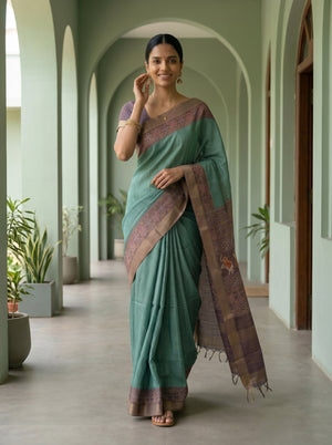Woman in a green saree with a purple border standing in a hallway with plants.
