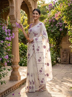 Woman in a white saree with purple floral patterns standing in a garden setting.