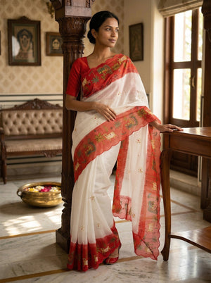 Woman in a white and red saree standing in an elegant room with decor.