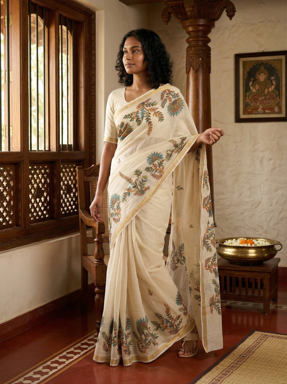 Woman in a cream saree with floral patterns standing in a traditional setting.
