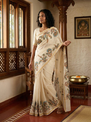 Woman in a cream saree with floral patterns standing in a traditional setting.