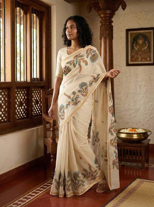 Woman in a cream saree with floral patterns standing in a traditional setting.