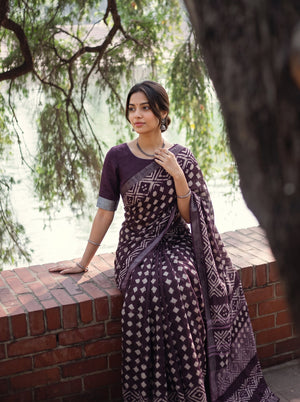 A woman wearing a dark purple and beige patterned saree leans against a brick wall outdoors near water and trees.