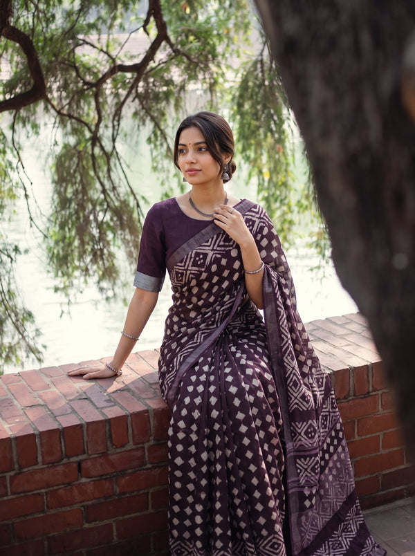 A woman wearing a dark purple and beige patterned saree leans against a brick wall outdoors near water and trees.