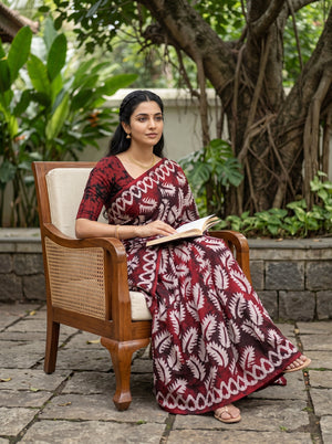 A woman sits in a wooden armchair outdoors, wearing a long-sleeved maroon and beige printed dress and holding an open book. The background features a stone wall, lush green plants, and a large tree.
