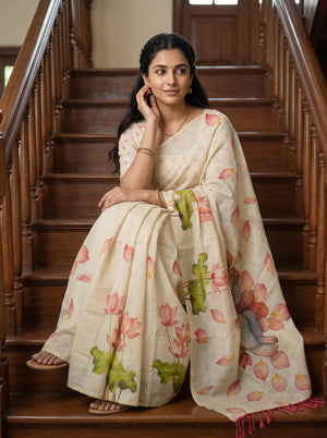 A woman sits on wooden stairs wearing a cream-colored saree with a pink and green lotus print. The saree has pink lotus flowers and green leaves, and the background consists of dark brown wooden stairs.