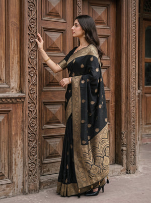 A woman wearing a black saree with gold motifs and a heavily embroidered gold border, standing in front of ornate carved wooden doors.