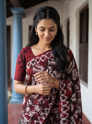 A woman wearing a dark red and white patterned saree, standing in a corridor with columns.