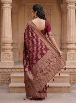 A woman wearing a deep maroon and gold Banarasi silk saree, viewed from behind as she stands in front of a traditional stone pillar structure.