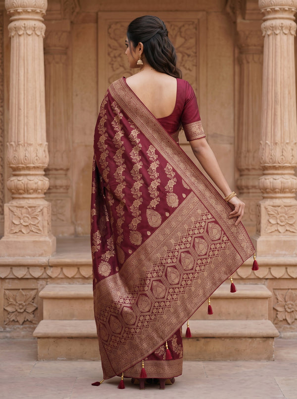 A woman wearing a deep maroon and gold Banarasi silk saree, viewed from behind as she stands in front of a traditional stone pillar structure.