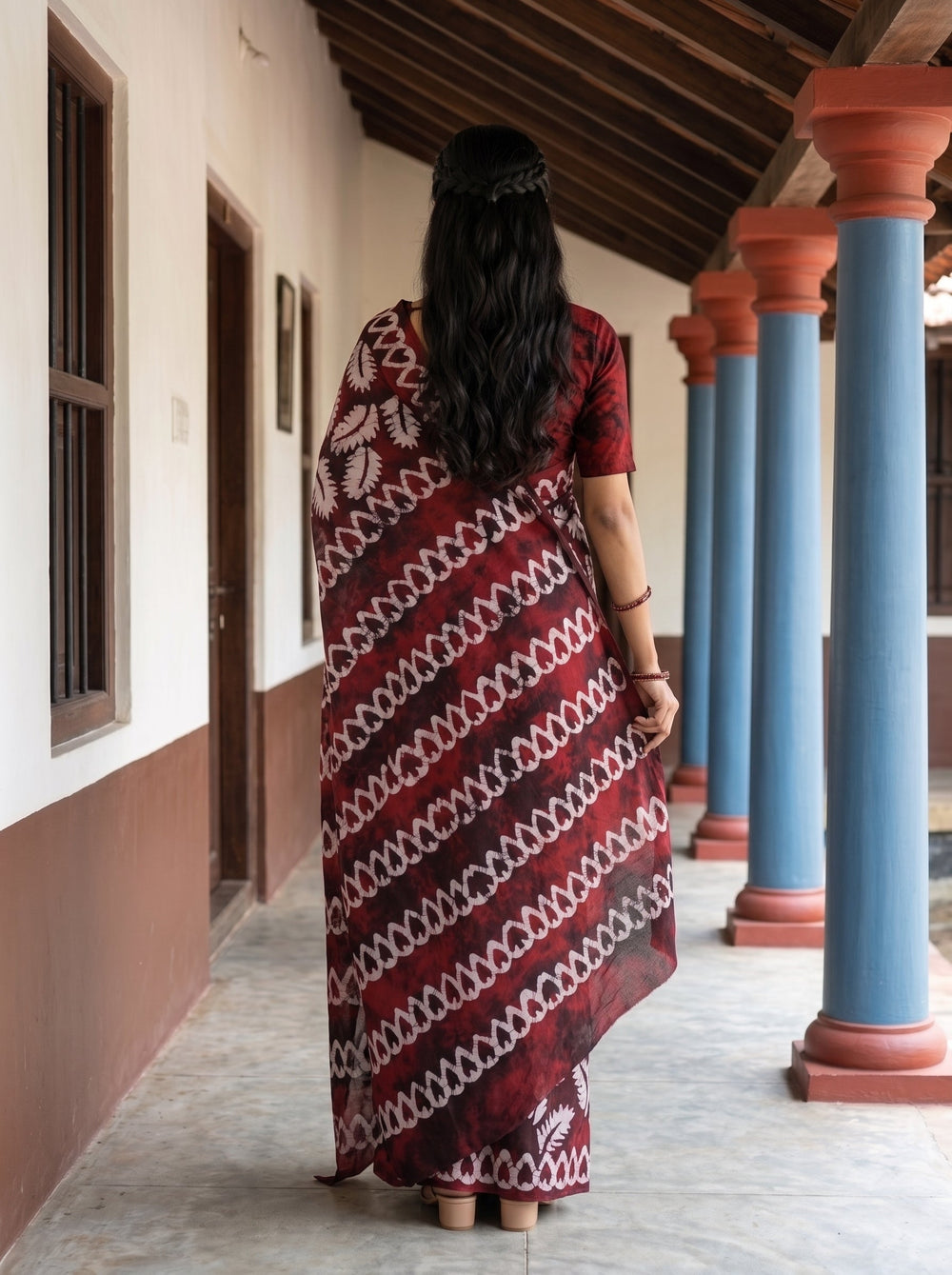 A woman wearing a dark red and white patterned silk saree stands in a traditional corridor with blue and terracotta pillars.