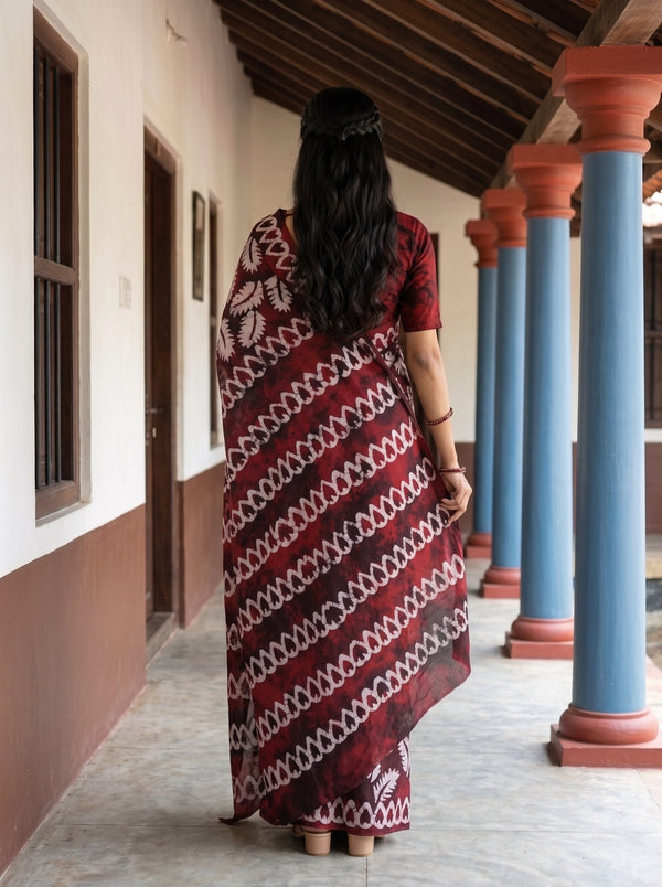 A woman viewed from behind wearing a red and white patterned sari while standing on a covered walkway with blue and terracotta columns.