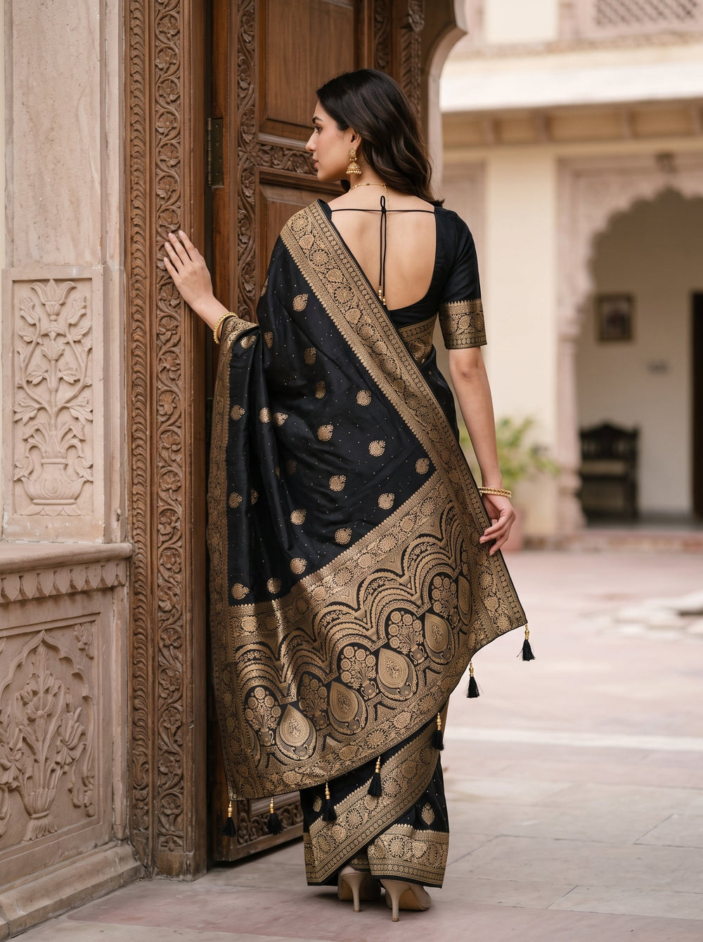 A woman wearing a black silk saree with intricate gold zari embroidery and a wide border, standing in front of a carved stone wall and wooden door.