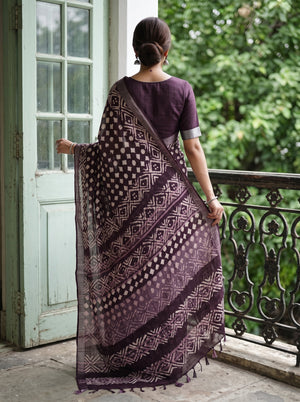 A woman stands on a balcony wearing a dark purple saree with a geometric block print pattern and a matching purple blouse, photographed from behind.