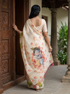 A woman viewed from behind wearing a cream-colored saree with a colorful illustration of a child and scattered pink lotus petals, standing in a traditional courtyard next to a carved wooden door.