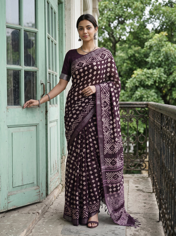 A woman stands on an outdoor balcony wearing a dark purple and beige patterned saree with a matching short-sleeve blouse. The background features a light green wooden door and lush green trees.