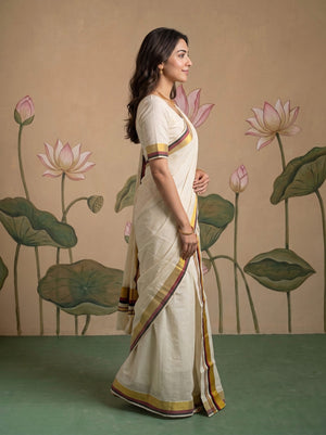 A woman wearing a cream-colored cotton saree with yellow, brown, and red striped borders, standing in profile against a beige wall with a painted lotus motif.