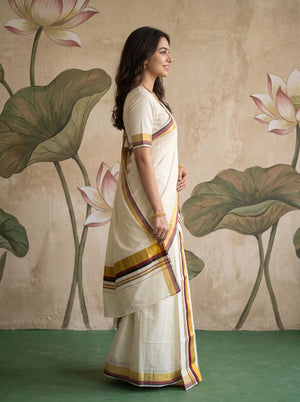 A woman wearing a cream-colored cotton saree with yellow, orange, and purple border stripes, standing in profile against a textured beige wall with painted lotus motifs.