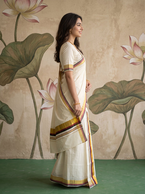 A woman wearing a cream-colored cotton saree with yellow, orange, and purple border stripes, standing in profile against a textured beige wall with painted lotus motifs.