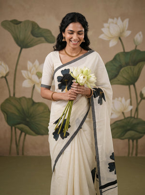 A woman wearing a cream-colored sari with black floral motifs and gray border details, holding a bouquet of white lotus flowers against a backdrop featuring a painted lotus pond mural.