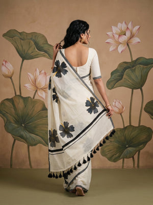 A woman wearing a white saree with black floral motifs and black tassels, shown from the back against a beige wall with a painted lotus mural.