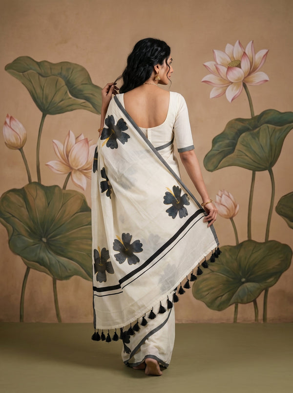 A woman wearing a white saree with black floral motifs and black tassels, shown from the back against a beige wall with a painted lotus mural.