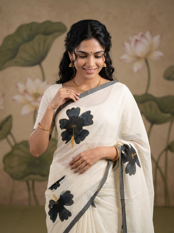 A woman wearing a white saree with black floral motifs and a gray border, standing against a beige background with lotus illustrations.