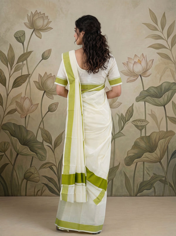 A woman wearing a white saree with lime green borders and a matching white blouse, shown from the back against a beige floral wall backdrop.