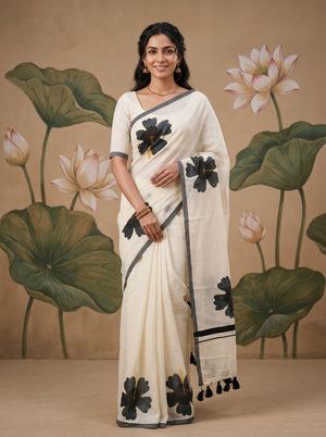 A woman models a cream-colored cotton saree with black floral prints and gray border details, standing in front of a beige backdrop decorated with painted lotus leaves and flowers.