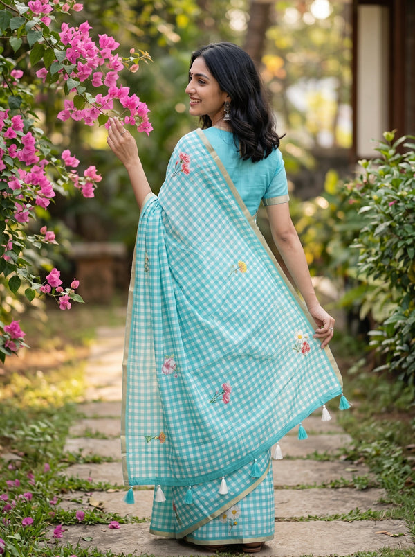 A woman wearing a light blue and white gingham saree with small floral embroidery and a gold border, standing on a garden path next to pink flowers.