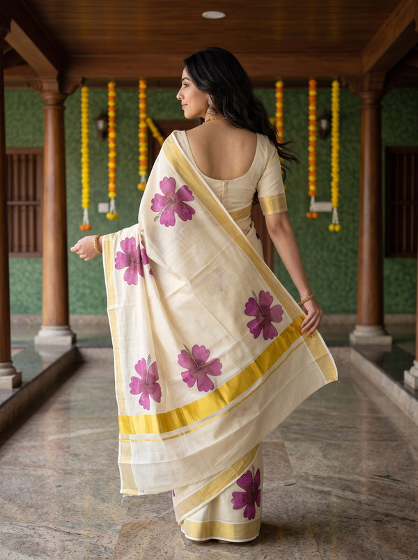 A woman wearing a cream-colored cotton saree with purple floral prints and yellow borders, standing in a traditional corridor with wooden pillars and green walls.