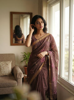 Woman in a brown saree standing by a window in a room with a couch and plant.