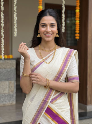 A woman wearing a white saree with purple and gold stripes, styled with gold jewelry, standing in front of a decorated doorway.