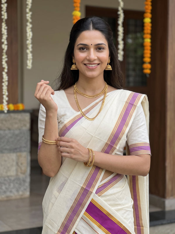 A woman wearing a white saree with purple and gold stripes, styled with gold jewelry, standing in front of a decorated doorway.