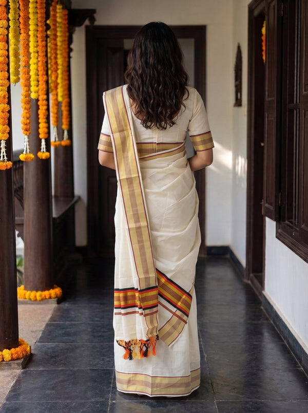 A woman viewed from behind wearing a cream-colored cotton saree with a beige and orange striped border, standing in a corridor decorated with yellow marigold garlands.