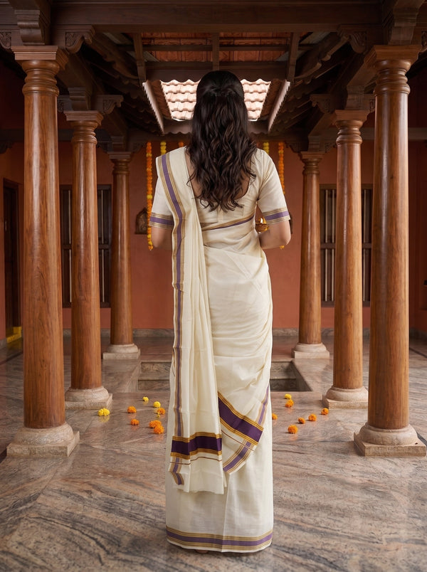 A woman viewed from behind wearing a cream-colored traditional saree with purple and gold border stripes, standing in a corridor with wooden pillars.