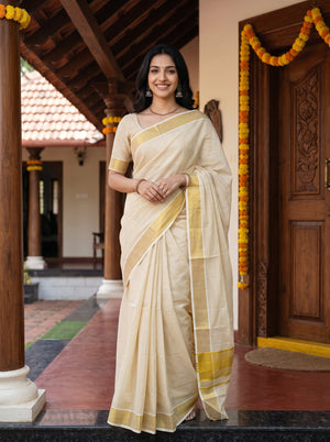 A woman wearing a light beige silk saree with yellow-gold borders stands on a traditional house porch decorated with marigold garlands.