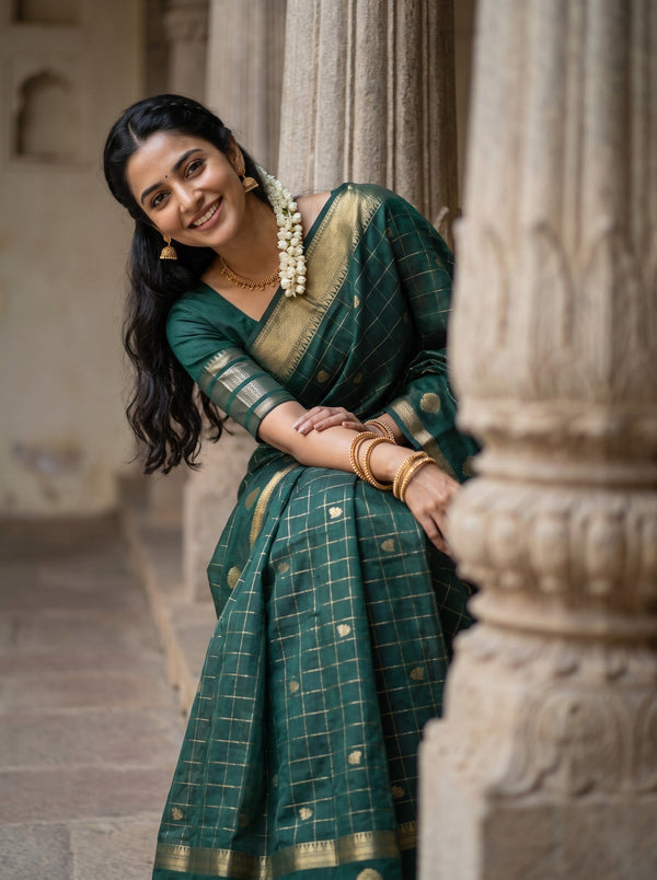 A woman wearing a dark green saree with gold grid patterns and a gold border, leaning against a stone pillar in an outdoor architectural setting.