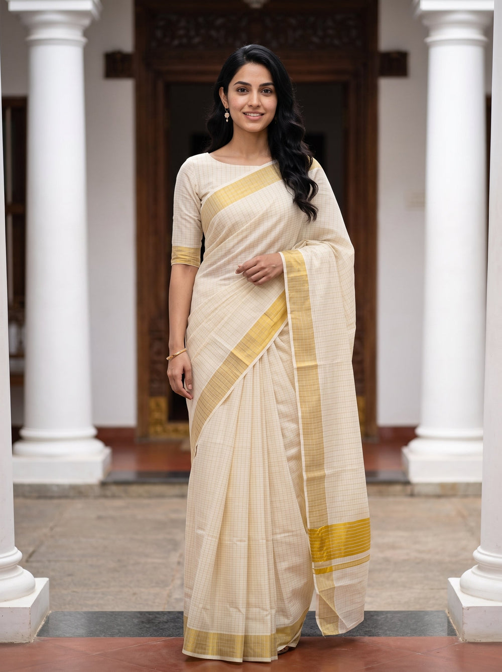A woman wearing a cream-colored cotton saree with a mustard yellow border and matching blouse, standing in a courtyard with white pillars.