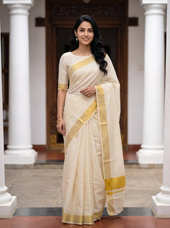 A woman wearing a cream-colored cotton saree with a mustard yellow border and matching blouse, standing in a courtyard with white pillars.