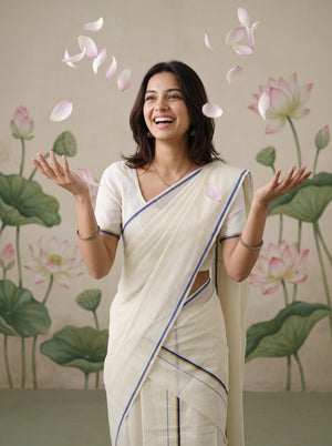 A woman wearing a cream-colored sari with blue and gold border stripes stands smiling with her hands raised, surrounded by floating pink lotus petals against a backdrop of painted lotus flowers.