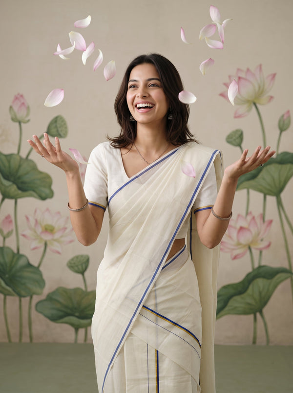 A woman wearing a cream-colored sari with blue and gold border stripes stands smiling with her hands raised, surrounded by floating pink lotus petals against a backdrop of painted lotus flowers.