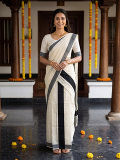 A woman wearing a cream-colored cotton saree with black and gray stripes, paired with a matching cream blouse, standing in a traditional courtyard decorated with marigold garlands.