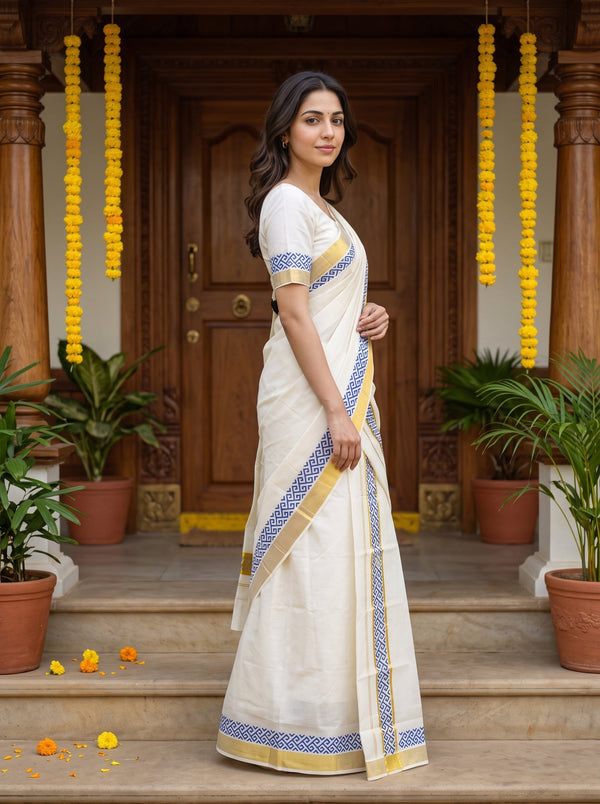 A woman wearing a white cotton saree with blue and gold borders, standing on stone steps in front of a wooden door decorated with yellow marigold garlands.